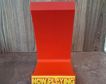 Puede incluir: Un soporte de exhibición de color rojo brillante con respaldo curvo y base horizontal. El borde frontal de la base presenta una placa rectangular amarilla con el texto rojo en relieve "NOW PLAYING" y una pequeña estrella roja entre las palabras.
