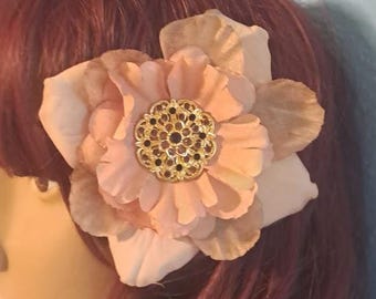 May include: A large, layered fabric flower hair accessory in shades of soft peach and light pink. The center of the flower features a round, intricate gold-tone metal embellishment with small, dark, circular accents arranged in a floral pattern.