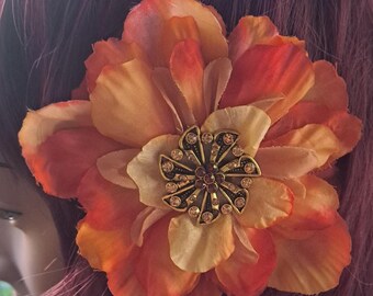 May include: A fabric flower hair accessory featuring layered petals in shades of orange, yellow, and red. The center of the flower is adorned with a circular, antique-gold-toned metal embellishment set with small, sparkling amber-colored crystals.