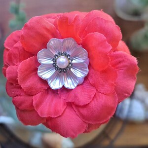 May include: A vibrant red fabric flower brooch featuring multiple layered petals. At the center, a white iridescent shell-like flower component is adorned with a small, round, cream-colored faux pearl surrounded by a ring of tiny, dark gray crystals.