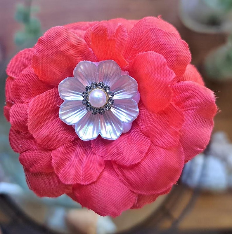 May include: A vibrant red fabric flower brooch featuring multiple layered petals. At the center, a white iridescent shell-like flower component is adorned with a small, round, cream-colored faux pearl surrounded by a ring of tiny, dark gray crystals.
