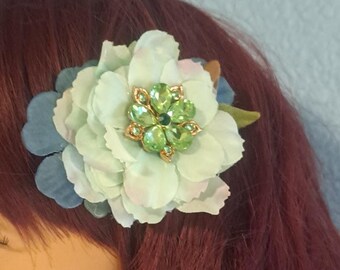 May include: A hair accessory featuring a layered fabric flower in shades of pale mint green and dusty blue. The center of the flower is adorned with a cluster of bright lime green faceted crystals set in a gold-toned metal frame.