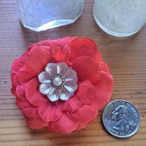 May include: A red fabric flower brooch featuring a central iridescent shell-like floral accent with a small pearl and dark crystal border. A United States quarter is placed next to the flower to provide a clear sense of scale for the object.