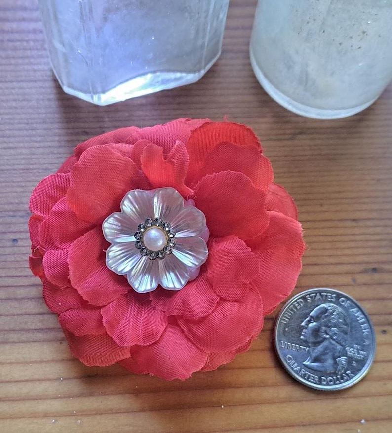 May include: A red fabric flower brooch featuring a central iridescent shell-like floral accent with a small pearl and dark crystal border. A United States quarter is placed next to the flower to provide a clear sense of scale for the object.