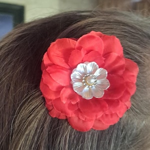 May include: A bright red fabric flower hair accessory featuring multiple layered petals. The center of the flower is adorned with a smaller, translucent white floral-shaped embellishment and a single central pearl-like bead.