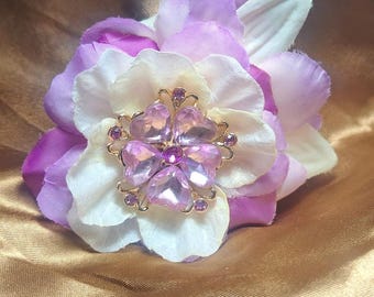 May include: A hair accessory featuring layered fabric flower petals in shades of white and purple. The center of the flower is adorned with a gold-toned metal embellishment shaped like a five-petaled blossom, set with pink heart-shaped crystals.