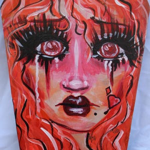 May include: A painted artwork featuring a stylized face with red hair and a coffin-shaped background. The face has large eyes with long black lashes, tears, and dark red lips. A heart and diamond are painted on the cheek.