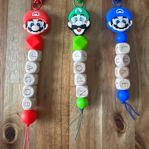 May include: Three character-themed keychains featuring Mario, Luigi, and a blue-hatted character. Each keychain has a plastic clip, a silicone character head, a geometric silicone bead, and wooden letter beads spelling Jacob, John, and Luke.