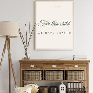 Op de afbeelding: Een ingelijste print met de tekst "For this child we have prayed" hangt boven een houten console. De tafel heeft lades, geweven manden en decoratieve items. Een vloerlamp en vaas met takken zijn ook zichtbaar.