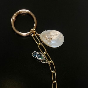 Op de afbeelding: Goudkleurige ketting sleutelhanger met schelp, schildpad en andere bedels. De sleutelhanger heeft een grote gouden ring, een witte schelp, een blauwe bedel, een witte schildpad bedel en een kleine schelp bedel.