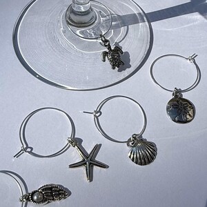 May include: A collection of silver-toned wine charms with various sea-themed pendants, including a turtle, starfish, seashell, and sand dollar. The charms are attached to small silver hoops, designed to hang on the rim of a wine glass. The background is a bright white surface.