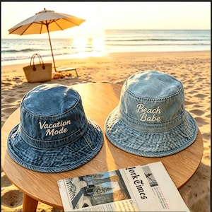 Puede incluir: Dos sombreros de pescador de mezclilla con texto bordado en blanco. Uno dice "Vacation Mode", el otro "Beach Babe". Los sombreros están sobre una mesa de madera con un periódico, una sombrilla de playa y una bolsa de paja en el fondo.