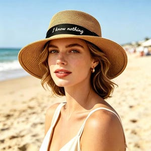 May include: A wide-brimmed straw sun hat with a black band bearing the text "I know nothing". The hat is a light tan colour and has a round crown. The background shows a beach scene with the sea and sky.