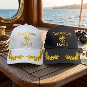 Op de afbeelding: Een witte honkbalpet en een zwarte honkbalpet, beide voorzien van goudkleurig borduurwerk met de tekst Commodore David, een gouden kompasroos-icoon en gouden laurierbladeren op de klep. Beide petten hebben een gestructureerde kroon.