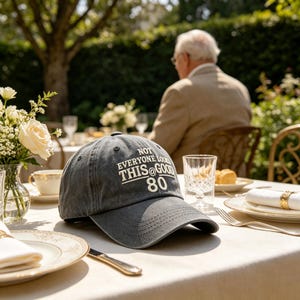 Puede incluir: Gorra de béisbol gris con el texto blanco "NOT EVERYONE LOOKS THIS @ GOOD 80". La gorra está sobre una mesa puesta para una comida, con una persona al fondo. La escena es al aire libre, con flores y vegetación.
