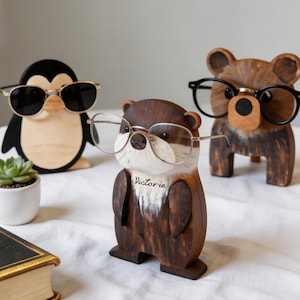 Puede incluir: Soportes de gafas de madera con forma de animales. La colección incluye un pingüino con gafas de sol, una nutria con gafas y un oso con gafas. La nutria tiene grabado el nombre "Victoria". Los soportes son marrones y beige.