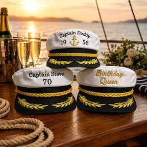 Könnte beinhalten: Drei weiß-schwarze Kapitänsmützen mit goldener Kordelverzierung und gestickten Lorbeerblättern auf dem Schirm. Die obere Mütze zeigt Captain Daddy 1956 mit einem Anker, die untere linke Captain Steve 70 und die untere rechte Birthday Queen.