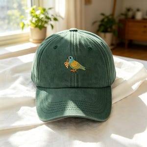 May include: A dark green baseball cap with a curved brim. The cap features an embroidered design of a colorful bird holding a slice of pizza in its beak. The cap is made of a soft, textured fabric.