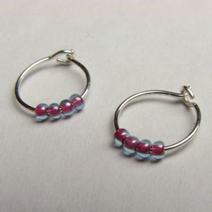 May include: A pair of small silver hoop earrings. Each earring features a row of five beads. The beads alternate between a translucent light blue and a deep pink color. The earrings are displayed on a light gray surface.