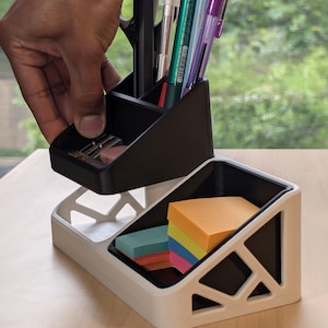 May include: A black and white desk organizer with a geometric design. The black section holds pens, scissors, and a pencil sharpener. The white section contains sticky notes. The organizer is made of plastic and is on a wooden surface.