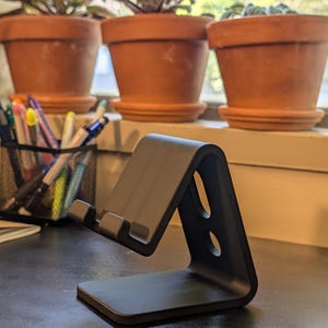 May include: A black, angled phone stand with two circular cutouts. The stand is on a dark surface, with three terracotta pots of plants in the background. A pen holder with pens and markers is also visible.