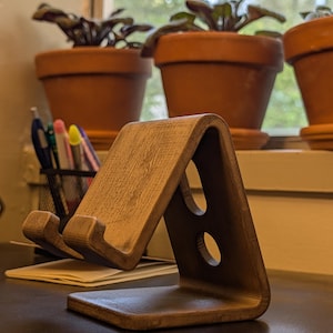 May include: A wooden phone stand with a light brown finish. The stand has a triangular shape with two circular cutouts and a ledge to hold a phone. The stand is on a dark surface, with potted plants in the background.
