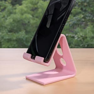 May include: A pink phone stand with a ribbed design holds a black smartphone. The stand has a triangular shape with a horizontal base and a vertical support, featuring two oval cutouts. The phone is angled for viewing.