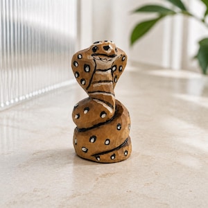 May include: A hand-carved wooden cobra figurine featuring a coiled body and an expanded hood. The light brown wood surface is decorated with dark brown and white painted spots, with black painted eyes and mouth details on the snake's head.