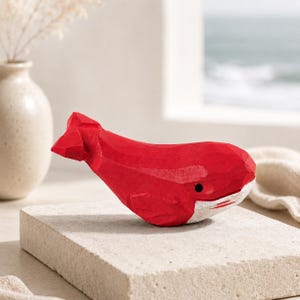 May include: A small, hand-carved wooden whale figurine painted in a vibrant red color. The whale features a faceted, geometric surface texture, a single black painted eye, and a white painted underside along the jawline and belly area.