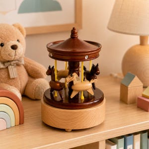 May include: A wooden carousel music box featuring a dark brown roof with small clear accents, supported by yellow poles. The carousel has three small carved horses in varying shades of brown and tan, mounted on a circular base atop a light wood stand.