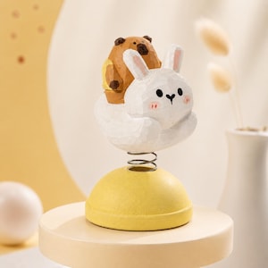 May include: A hand-carved wooden figurine featuring a white rabbit with pink cheeks and a brown bear sitting on its back. The figures are attached to a yellow semi-spherical base by a metal spring, allowing the top portion to bobble and move.