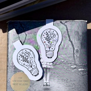 May include: Two white and black lightbulb-shaped bookmarks with floral designs and star accents. One bookmark is placed on a book with the text "THE NEW YORK TIMES BEST SELLER" on a gold sticker. The background is a blurred outdoor scene.