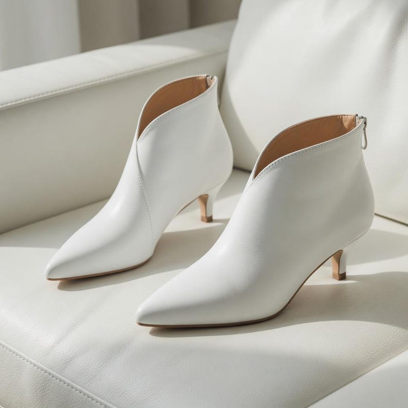 May include: A pair of white leather ankle boots with a pointed toe and a low, kitten heel. The boots have a V-shaped cut at the ankle and a zipper closure at the back. The heels measure approximately 2 inches.