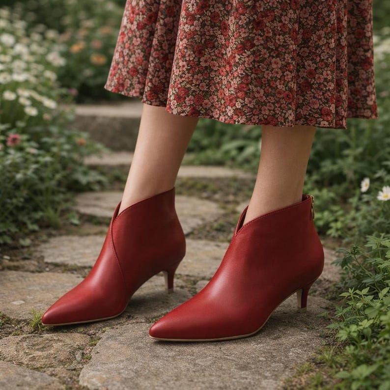 May include: Red leather ankle boots with a pointed toe and a low heel. The boots have a V-shaped cut at the ankle and a small zipper detail on the back. The boots are worn outdoors on a stone path.
