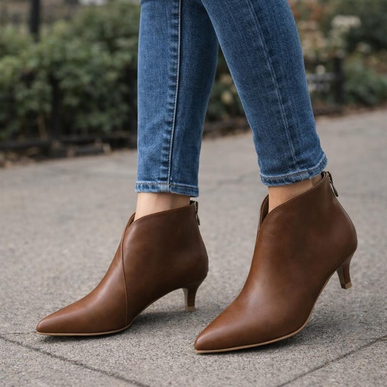 May include: Brown ankle boots with a pointed toe and a low heel. The boots have a V-shaped cutout on the front and a zipper on the back. The boots are worn with blue jeans.