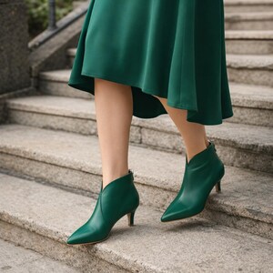 May include: Emerald green ankle boots with a pointed toe and a low heel. The boots have a V-shaped cut at the ankle and a zipper on the back. The boots are worn with a green dress, and the person is walking down a set of stone steps.