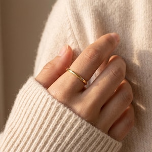 Puede incluir: Un anillo de banda de oro pulido con una superficie lisa y redondeada. El anillo presenta un diseño circular continuo y minimalista con un acabado metálico de alto brillo que refleja la luz, creando un brillo cálido sobre el metal.