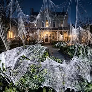 May include: A residential house exterior decorated with white synthetic spider webs draped over green bushes and trees. The two-storey building features illuminated windows and a front porch, creating a layered effect with the white webbing.