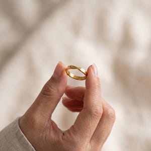 Op de afbeelding: Een goudkleurige ring met een gladde, gepolijste afwerking en een subtiel gedraaid designelement aan de bovenkant. De ring is cirkelvormig met een consistente breedte en heeft een reflecterend metalen oppervlak dat licht vangt.