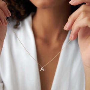 Puede incluir: Un collar de cadena fina en tono dorado con un pequeño colgante en forma de letra A mayúscula. El colgante de letra está incrustado con pequeñas piedras claras y brillantes que captan la luz, suspendido de una cadena de eslabones metálicos.