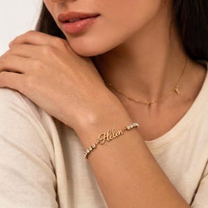 Puede incluir: Una pulsera de tono dorado con una placa central que muestra el texto Helen en letra cursiva, flanqueada por una serie de pequeñas piedras claras de corte redondo en una delicada cadena metálica.