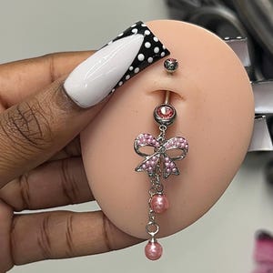 May include: A belly button ring with a pink bow and dangling pink pearls. The ring is silver-toned with a pink gem at the top and bottom. The bow is adorned with small pink gems. The image is of a belly button on a mannequin.