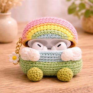 May include: A crochet case for wireless earbuds featuring a rainbow top in pink, yellow, and light blue. The base is light green with two white clouds and two small green spheres at the bottom. A gold chain with a white daisy charm is attached.
