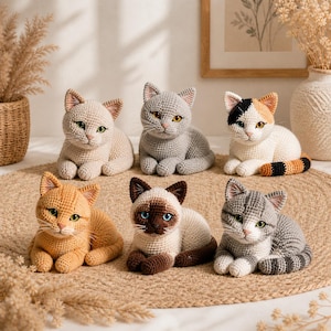 May include: Six crocheted cat ornaments in diverse colours, including beige, grey, orange, and Siamese patterns. Each cat has detailed features, such as eyes and markings, and is posed lying down. They are arranged on a round, woven mat.