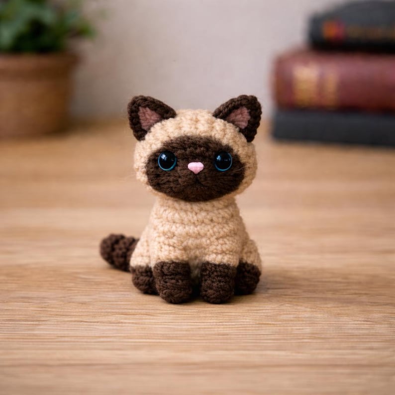 May include: A handmade crochet Siamese cat figurine. The cat has a tan body, dark brown face, ears, paws, and tail. It features large blue eyes and a pink nose. The cat is sitting on a wooden surface.
