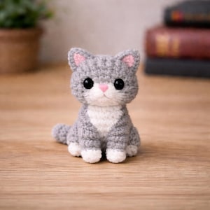 May include: A handmade gray and white crocheted cat figurine with pink accents on the ears and nose. The cat has large black eyes and is sitting upright. The figurine is on a wooden surface.