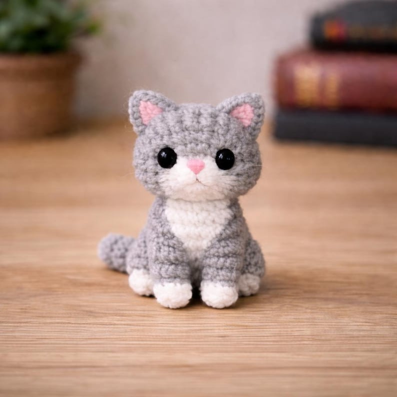 May include: A handmade gray and white crocheted cat figurine with pink accents on the ears and nose. The cat has large black eyes and is sitting upright. The figurine is on a wooden surface.