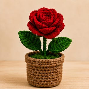 Puede incluir: Una rosa roja tejida a ganchillo en una maceta marrón. La flor hecha a mano tiene hojas verdes y está plantada en una maceta marrón tejida a ganchillo llena de hilo verde. El artículo decorativo es una artesanía única.