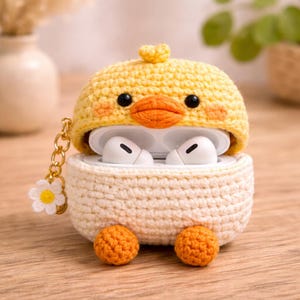 May include: A crochet case for wireless earbuds featuring a yellow duck design with a bright orange beak, black eyes, and two orange feet at the base. The bottom section is cream-coloured, and a gold-toned chain with a small white flower charm is attached.