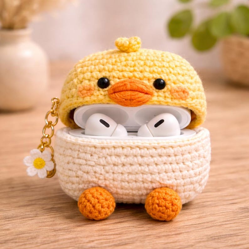 May include: A crochet case for wireless earbuds featuring a yellow duck design with a bright orange beak, black eyes, and two orange feet at the base. The bottom section is cream-coloured, and a gold-toned chain with a small white flower charm is attached.